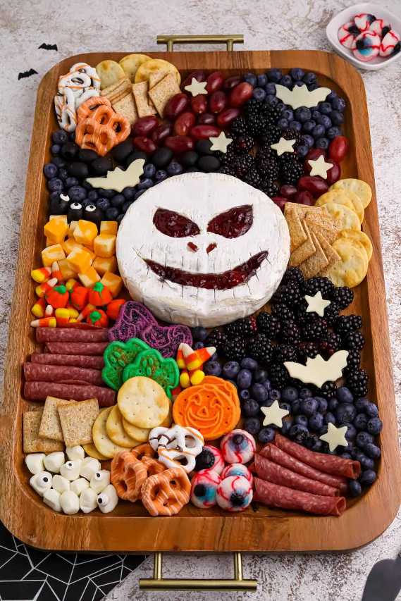 Dark wood board loaded with sliced meats, orange cheese wedges, gummy worms, and pumpkin-shaped crackers