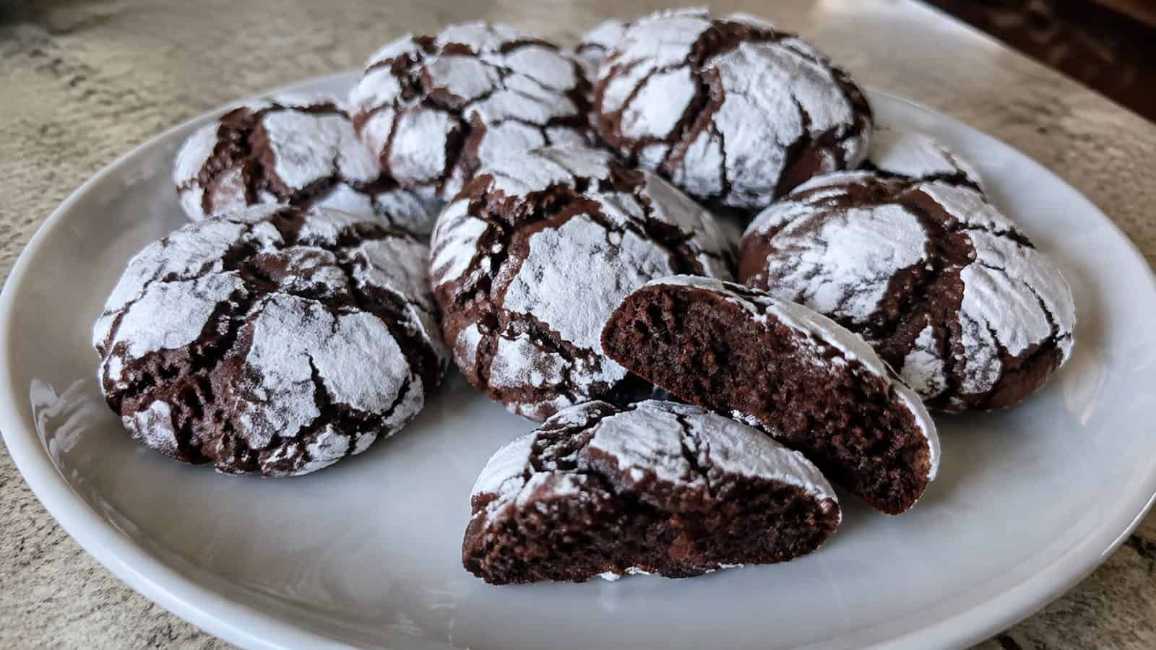 Dark chocolate cookies with a cracked white powdered sugar top on a dark slate board.
