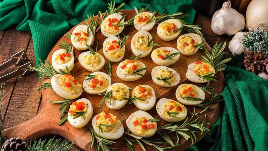 Halved deviled eggs topped with rosemary sprigs and diced red pepper confetti